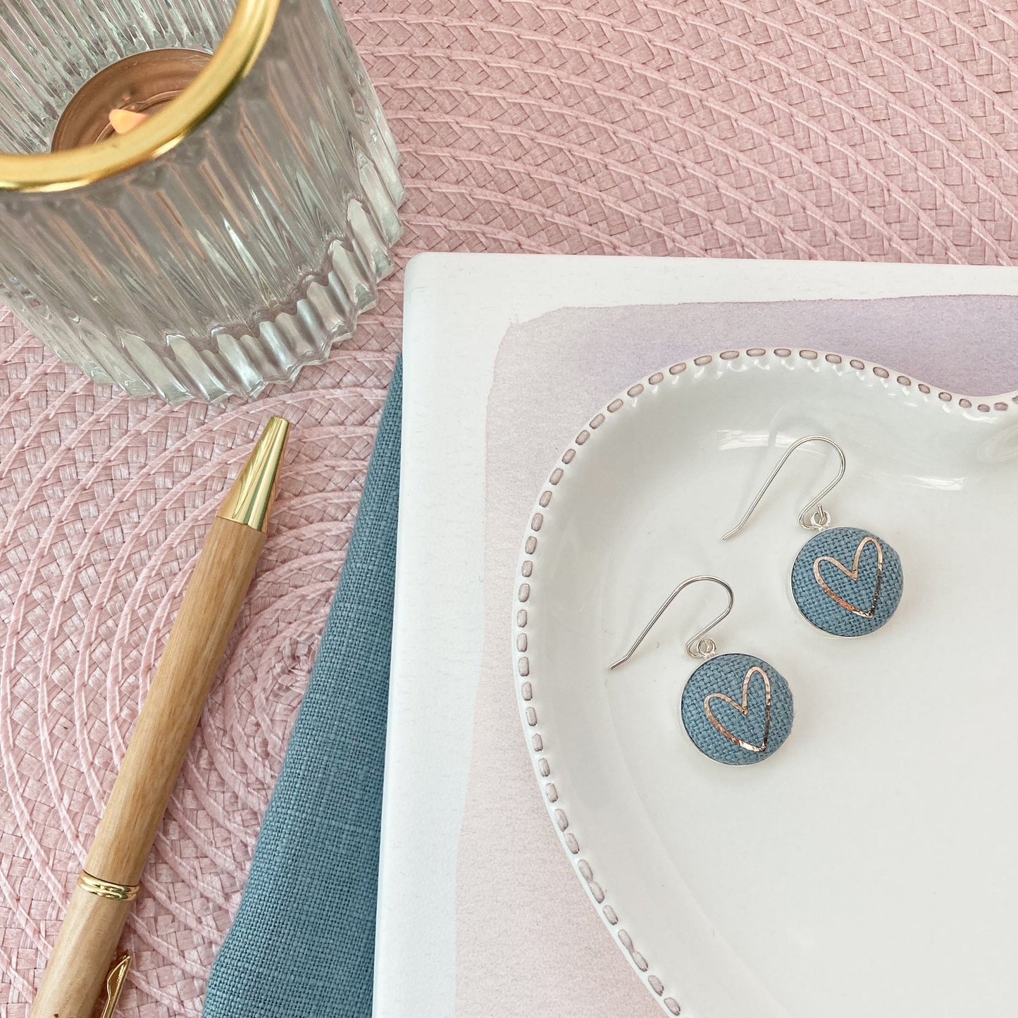 Blue heart-shaped earrings on a white dish with a pen and notebook on a pink textured surface.