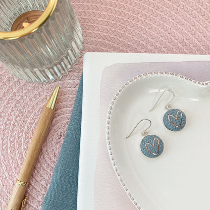 Blue heart-shaped earrings on a white dish with a pen and notebook on a pink textured surface.