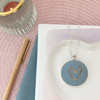 Necklace with heart pendant on a pink surface with a pen and notebook.