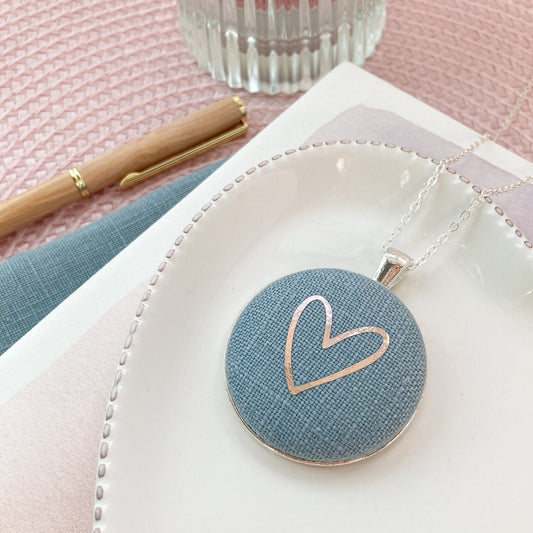 Round pendant with heart design on a white dish, next to a pen and glass on a pink surface.