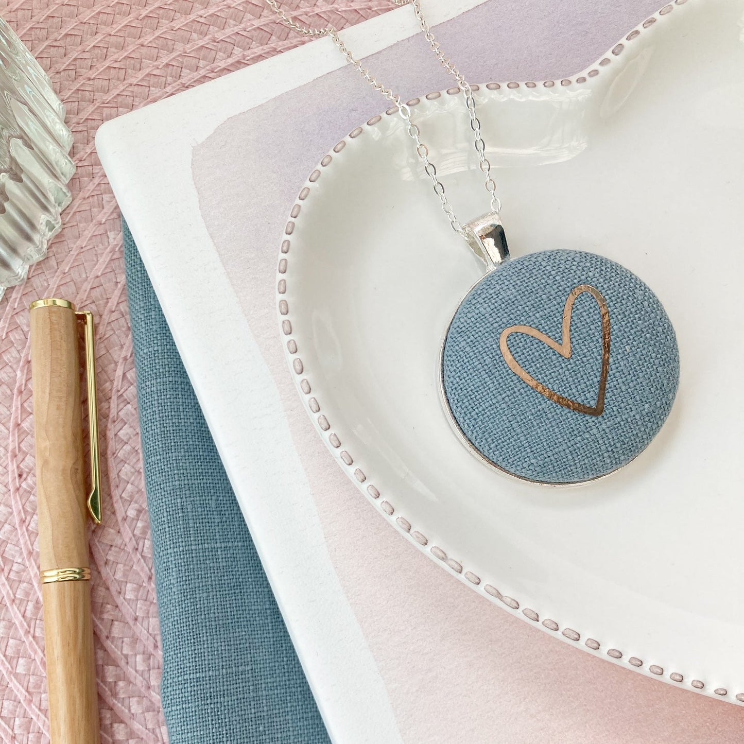Round pendant with heart design on a white dish, next to a pen and notebook on a pink and blue gradient background.