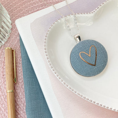 Round pendant with heart design on a white dish, next to a pen and notebook on a pink and blue gradient background.