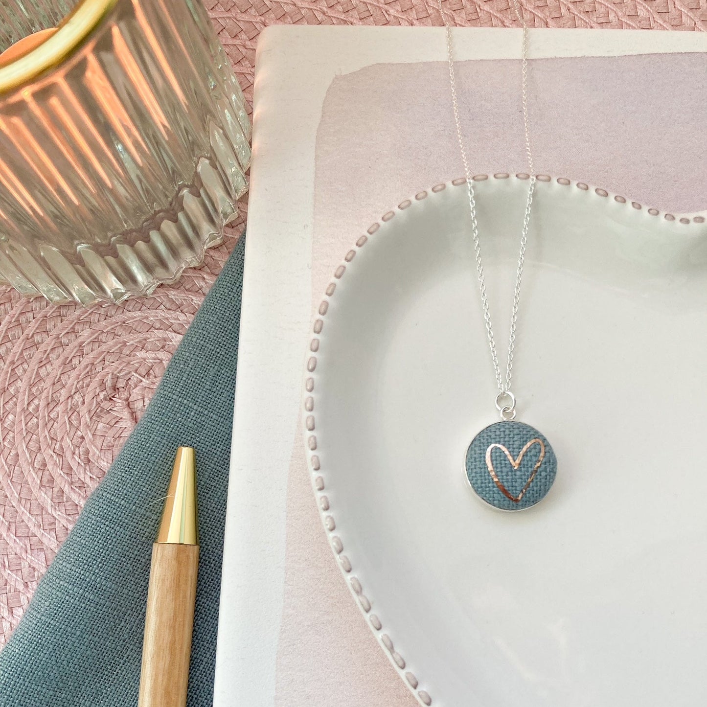 Necklace with a heart pendant on a pink surface with a pen and decorative item.