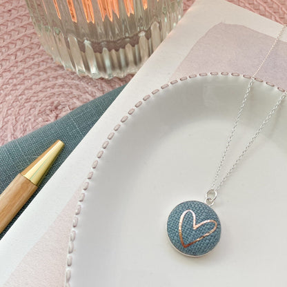 Grey blue heart necklace on a pink surface with a candle and pen.