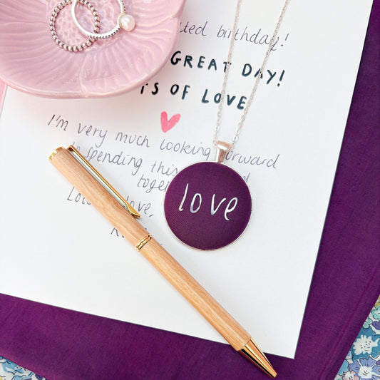 Round aubergine coloured fabric pendant necklace with handwritten "love" printed in silver, thread on a silver chain, placed on a handwritten birthday card and on top of aubergine & floral fabrics