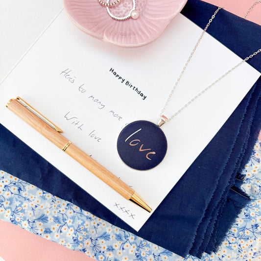 Navy fabric necklace pendant with the word love printed in silver handwriting, thread on a silver chain and placed on a handwritten card and navy fabric with a pen and pink bowl