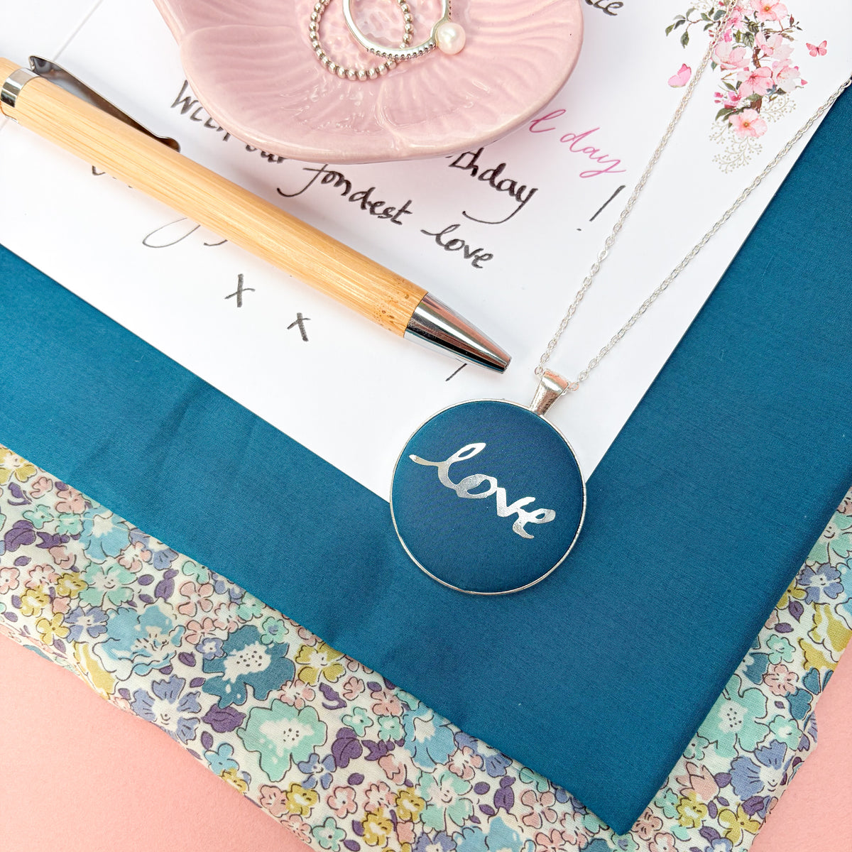 A blue fabric pendant necklace with the handwritten word "love" in silver print, thread on a silver chain and placed on a handwritten birthday card with fabrics and a pink bowl of rings
