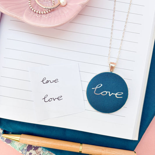 A blue fabric pendant necklace with the handwritten word "love" in silver print, thread on a silver chain and placed on a notebook with fabrics and a pink bowl of rings