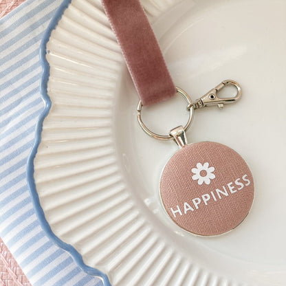 Keychain with 'Happiness' and a flower printed on pink blush fabric, on a decorative plate