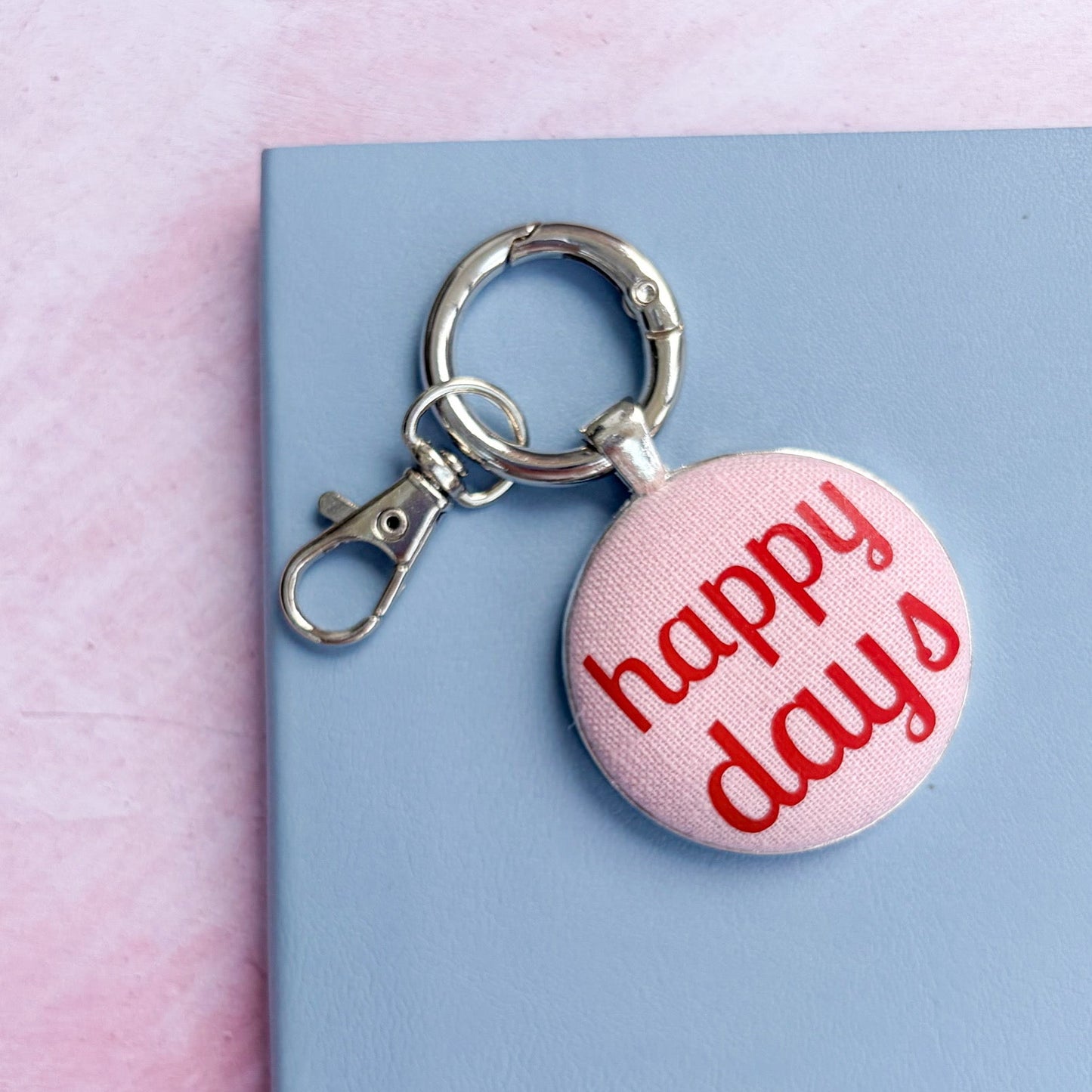 Keychain with 'happy days' text printed onto pink fabric, on a pink and blue background