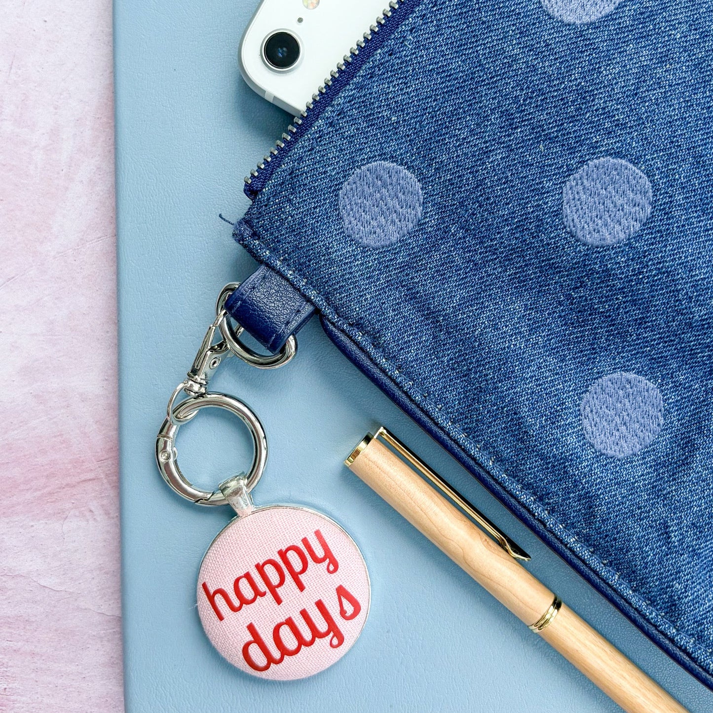 Blue polka dot pouch with a phone, pen, and 'happy days' keyring on a light pink background