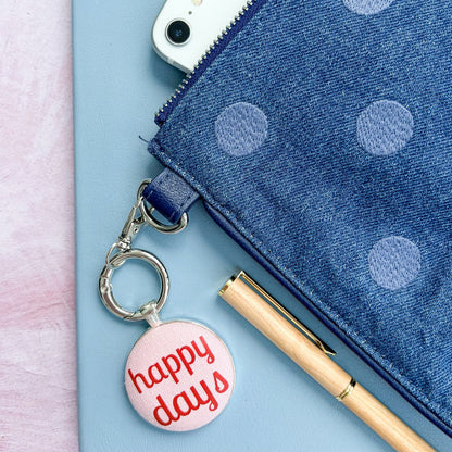 Blue polka dot pouch with a phone, pen, and 'happy days' keyring on a light pink background