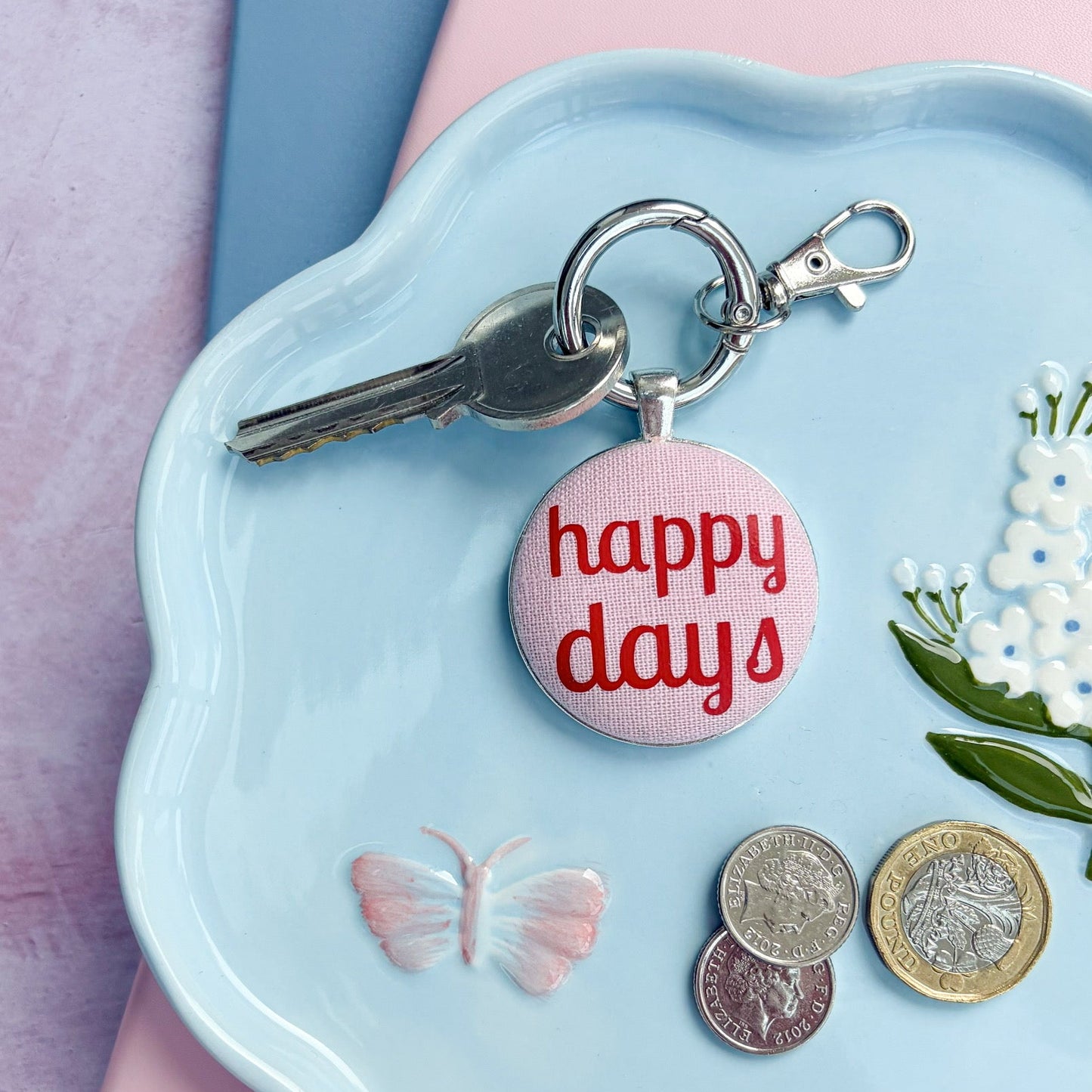 Keyring with 'happy days' charm on a decorative plate with coins and set on a pink and blue notebook