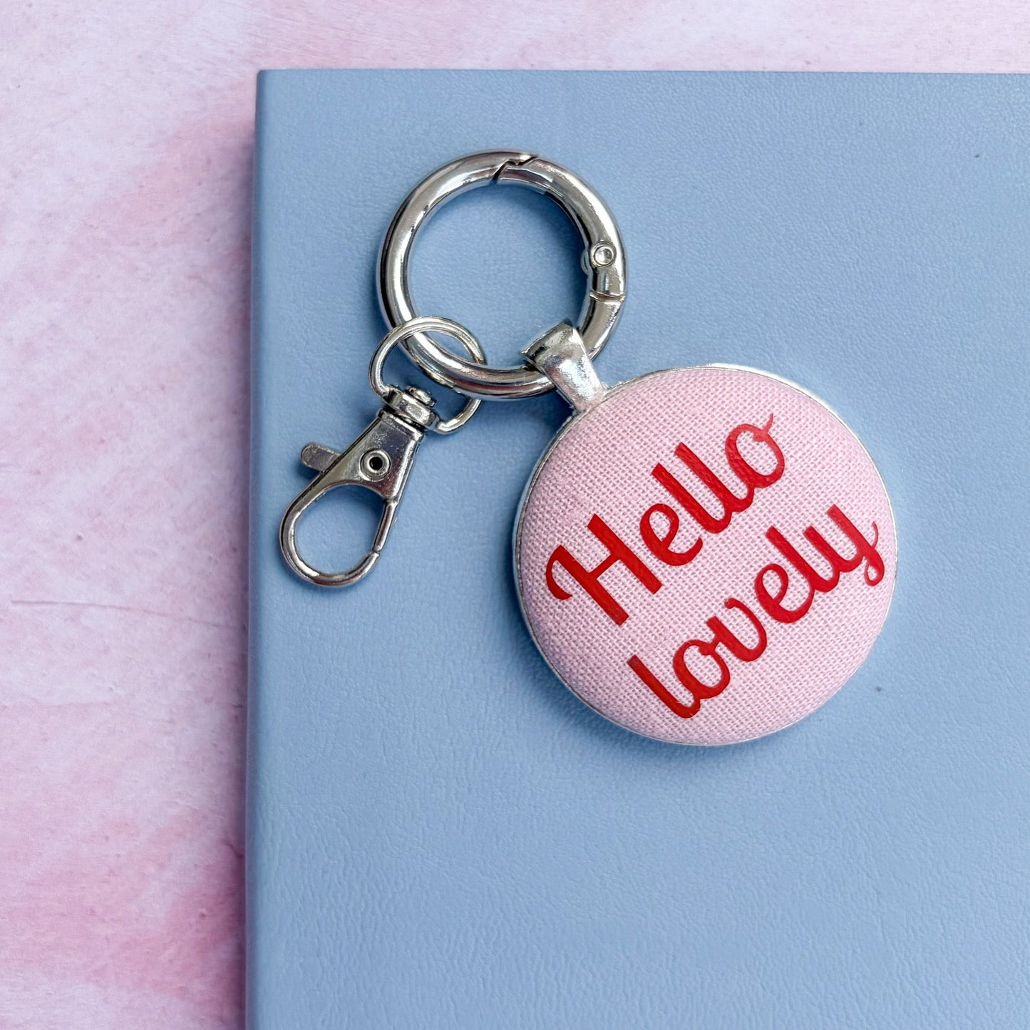 Keyring with 'Hello lovely' text printed in red on pink fabric, set on a pink and blue background