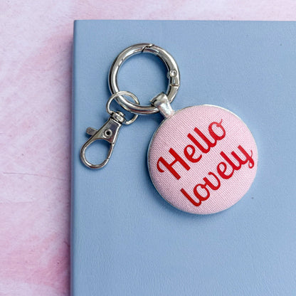 Keyring with 'Hello lovely' text printed in red on pink fabric, set on a pink and blue background
