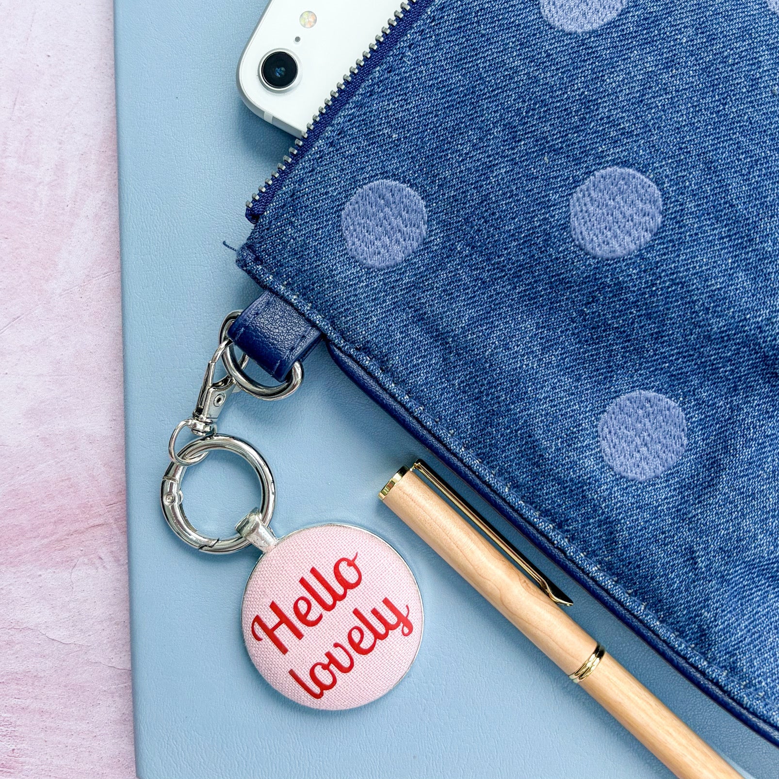 Keyring with 'Hello lovely' charm on a blue polka dot pouch with a phone inside, on a light pink background.