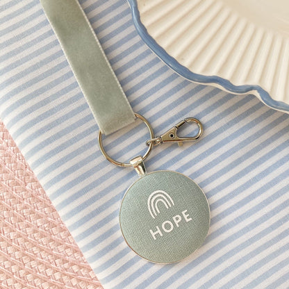 Key ring with 'HOPE' text and rainbow design on duck egg blue fabric, laid on a striped fabric background