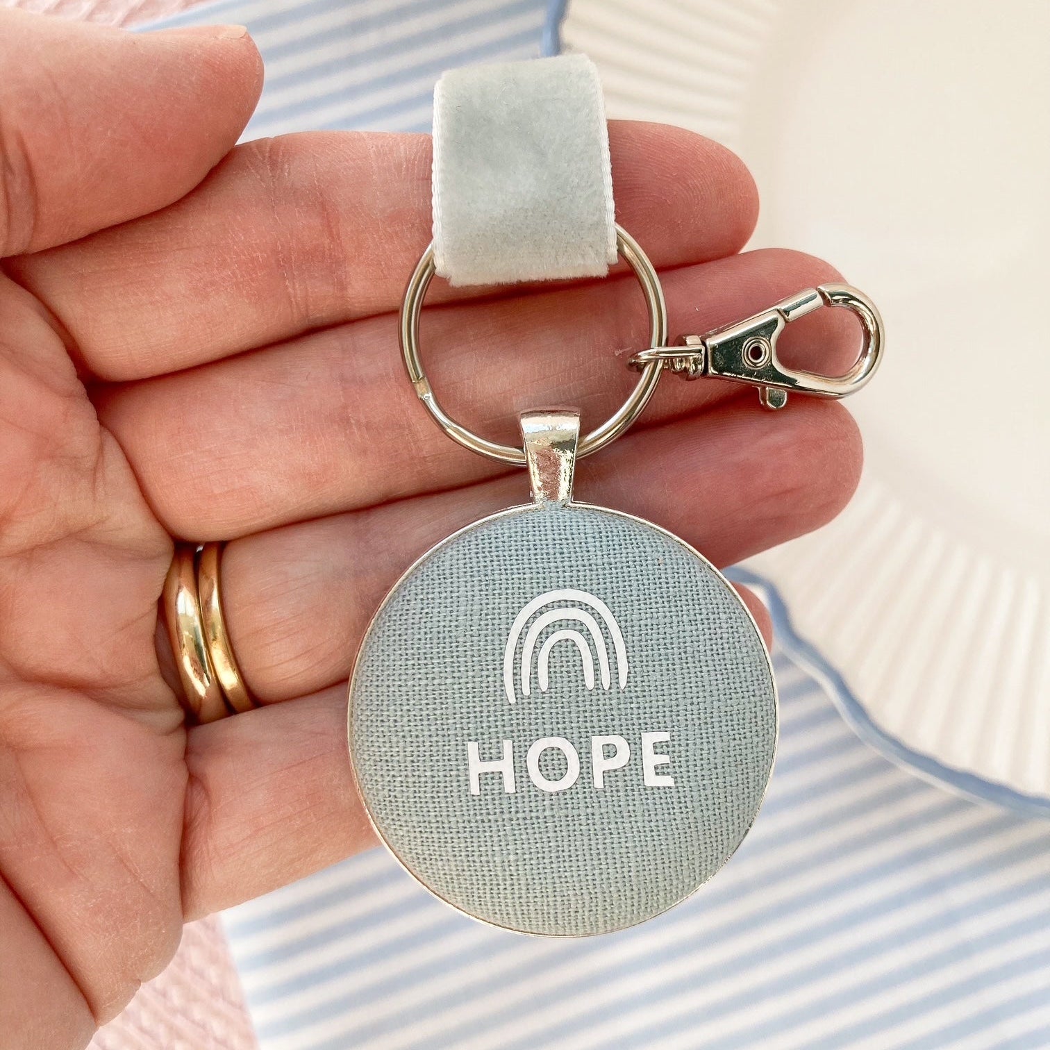 Keychain with 'HOPE' and rainbow symbol held in a hand against a striped fabric background