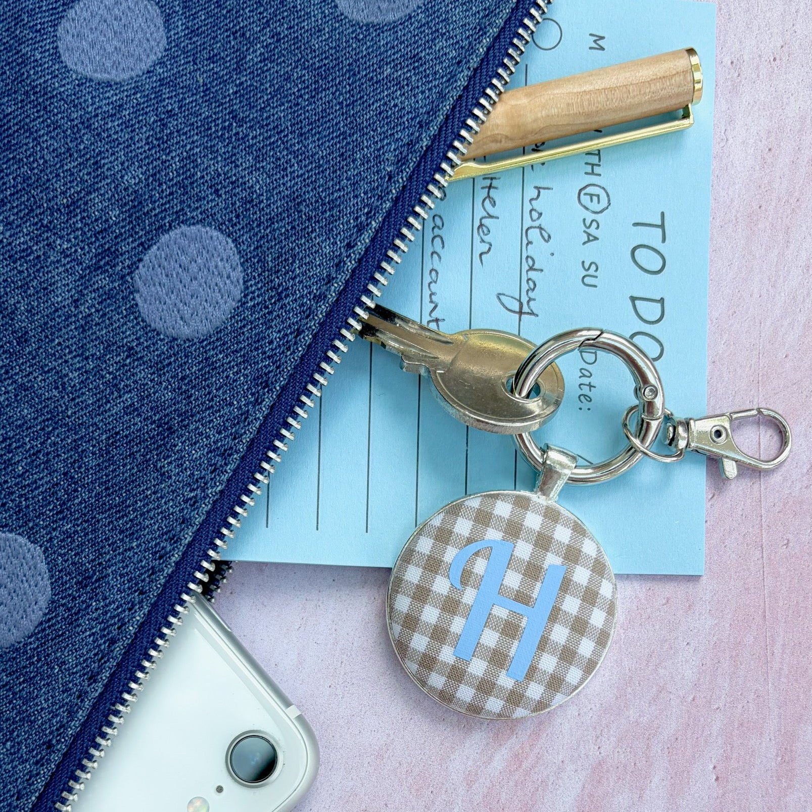 Caramel gingham fabric keyring with a blue H initial next to a blue denim clutch bag, pen and phone