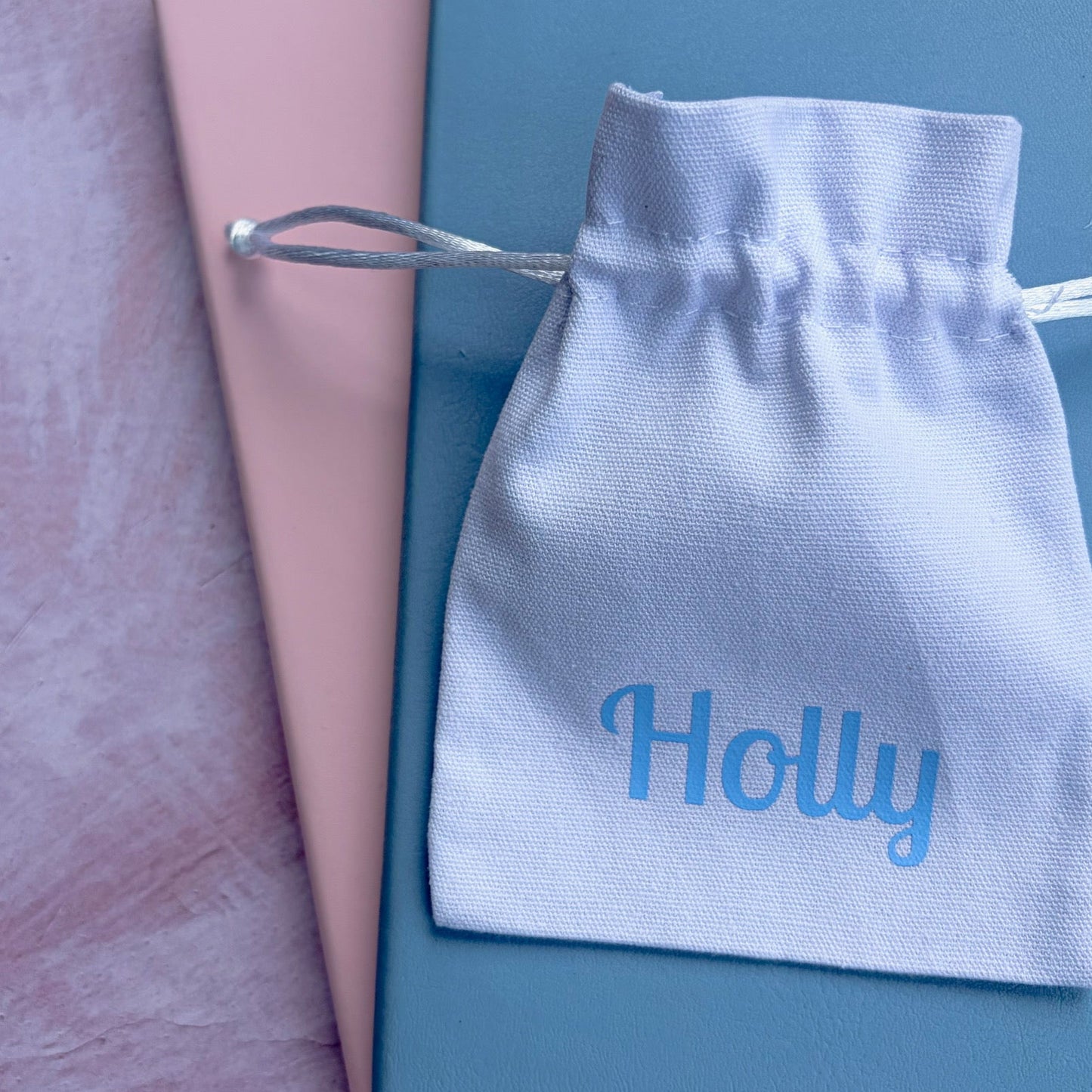 White drawstring gift pouch with 'Holly' printed in blue, on a pink and blue notebook