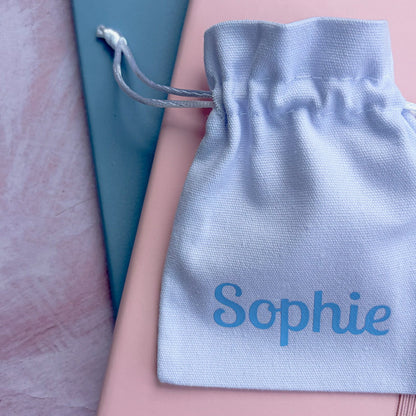 A white personalized drawstring pouch printed with the name 'Sophie' in turquoise blue, resting on top of a pink notebook and a teal book on a pink textured background.