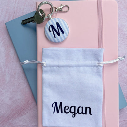 A white drawstring pouch printed with the name 'Megan' and a round striped monogram keychain with a silver clasp. The notebook is styled on top of a blue notebook against a textured pink background.