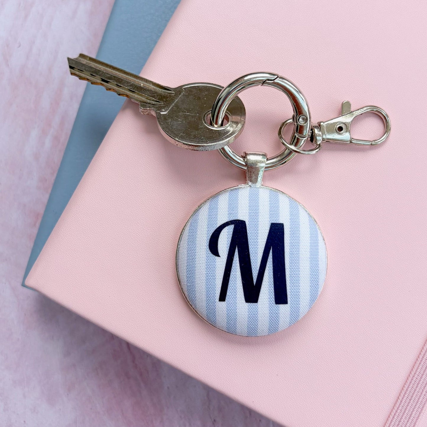 A personalized keychain with a round pendant featuring a navy blue monogram letter 'M' on a light blue and white striped background, attached to a silver keyring with a key and clasp, resting on a pink notebook.