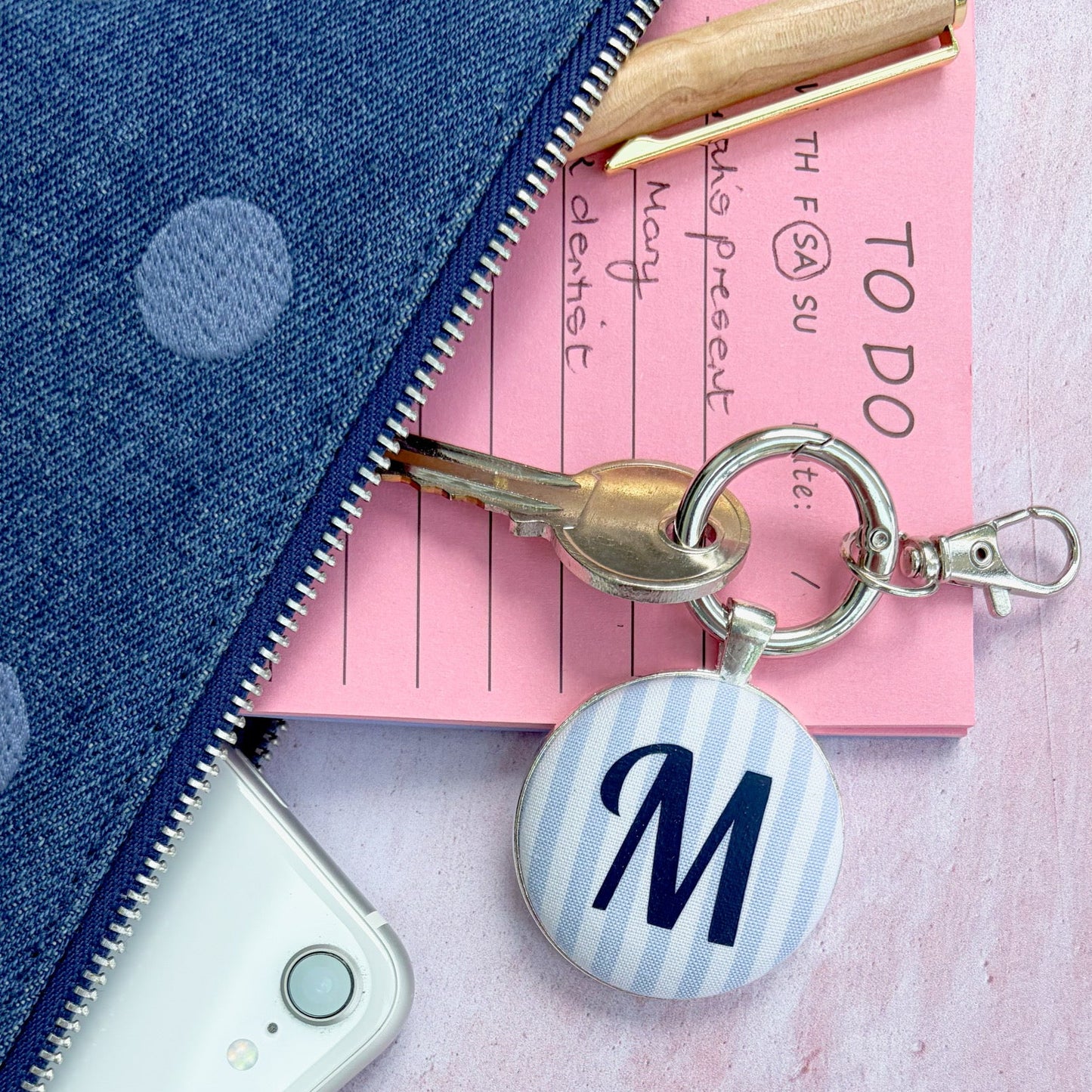 A personalized keyringwith a round button charm featuring a navy blue letter 'M' on a light blue and white striped background, attached to a silver key ring. The keychain is styled in a flat-lay arrangement alongside a pink notepad, a white smartphone, and a blue polka-dot zipper pouch on a textured pink surface.