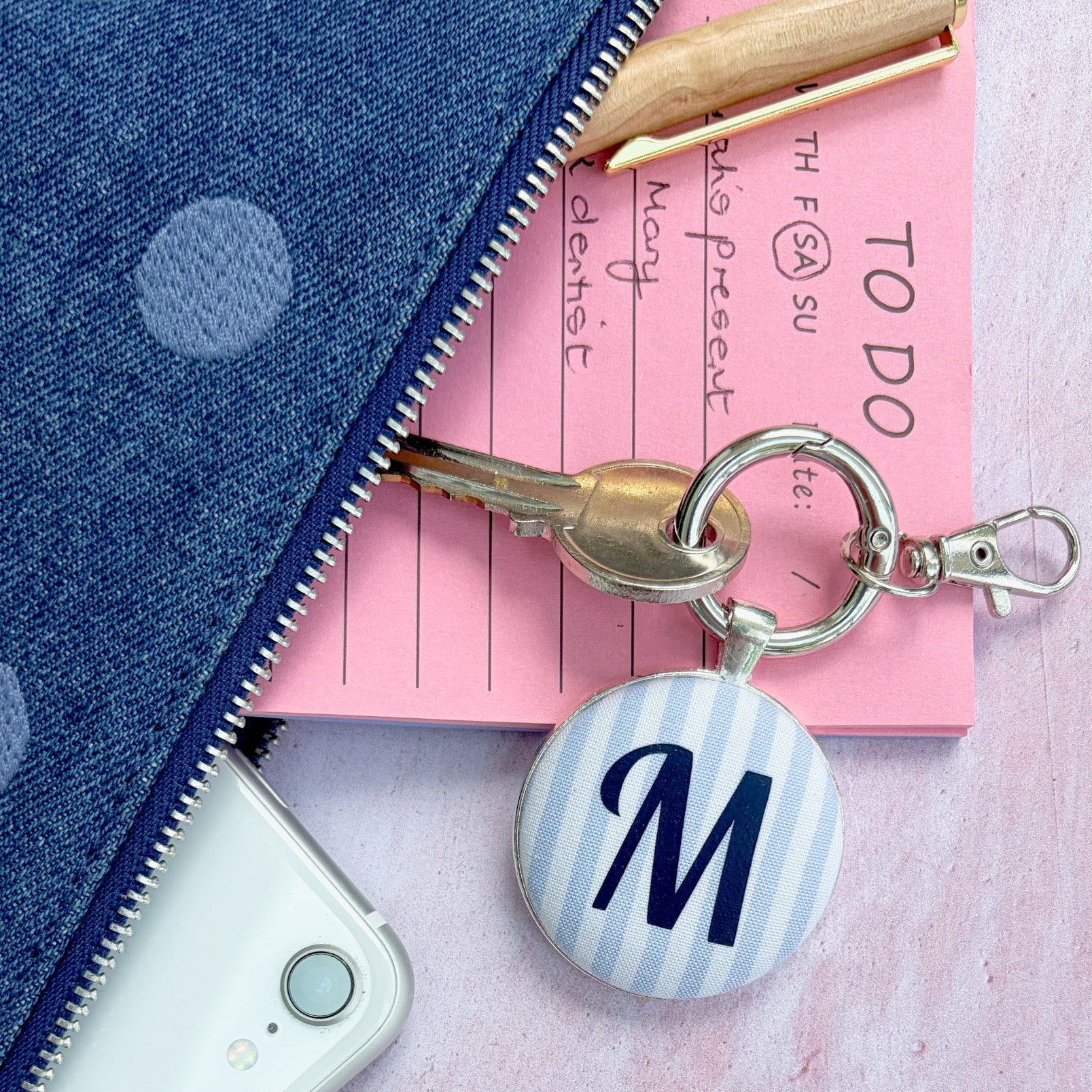 A personalized keyringwith a round button charm featuring a navy blue letter 'M' on a light blue and white striped background, attached to a silver key ring. The keychain is styled in a flat-lay arrangement alongside a pink notepad, a white smartphone, and a blue polka-dot zipper pouch on a textured pink surface.