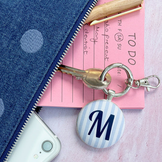 A personalized keyringwith a round button charm featuring a navy blue letter 'M' on a light blue and white striped background, attached to a silver key ring. The keychain is styled in a flat-lay arrangement alongside a pink notepad, a white smartphone, and a blue polka-dot zipper pouch on a textured pink surface.