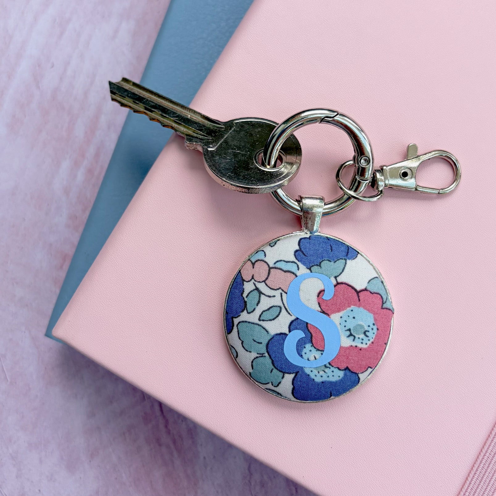 Keyring made with floral fabric and printed with a blue S initial, on a pink and blue notebook