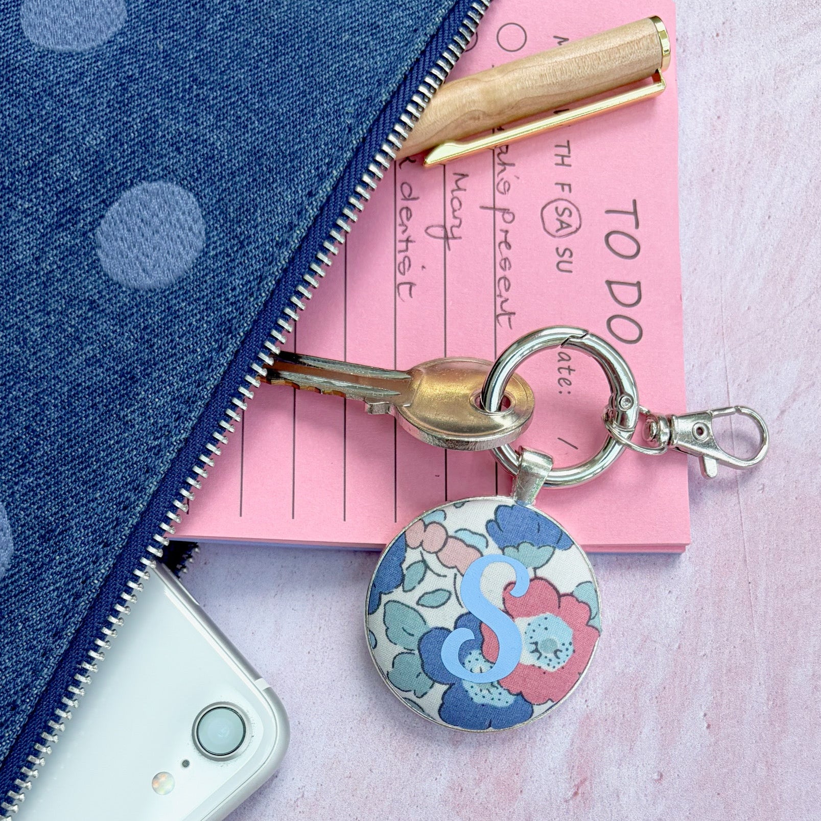 Floral fabric keyring with a blue S initial, next to a blue denim bag, phone and pen