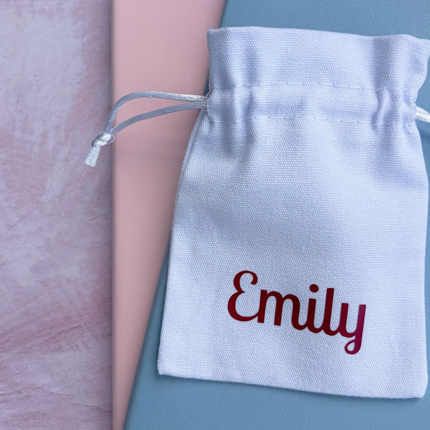 A personalized white cotton drawstring pouch embroidered with the name 'Emily' in burgundy, resting on top of a dusty blue notebook and a pink book on a textured pink surface.