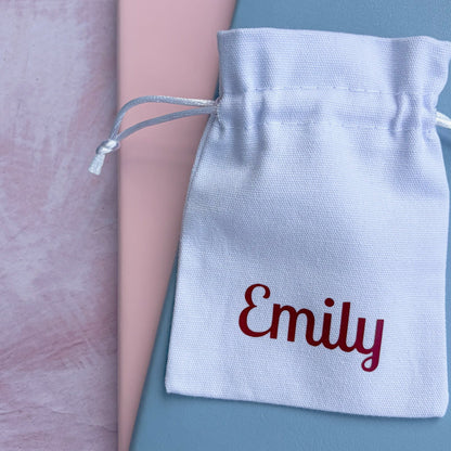 A personalized white cotton drawstring pouch embroidered with the name 'Emily' in burgundy, resting on top of a dusty blue notebook and a pink book on a textured pink surface.