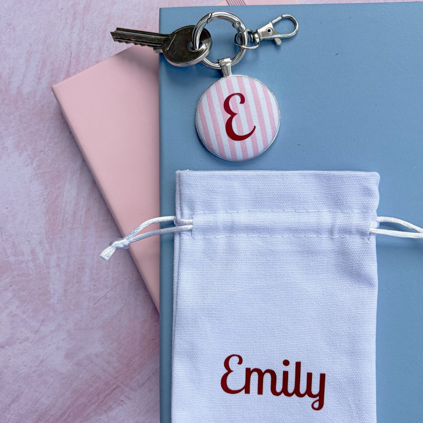 A personalized key ring featuring a silver metal key, a round pink and white striped button charm with a burgundy monogram 'E', and a white drawstring pouch embroidered with 'Emily' in burgundy, displayed on a light blue notebook over a pink background.