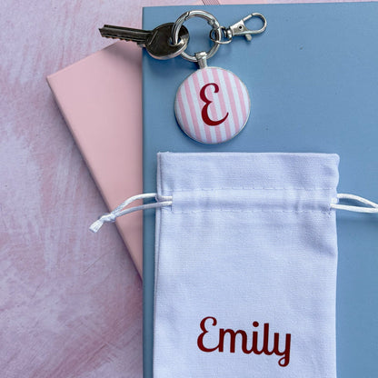 A personalized key ring featuring a silver metal key, a round pink and white striped button charm with a burgundy monogram 'E', and a white drawstring pouch embroidered with 'Emily' in burgundy, displayed on a light blue notebook over a pink background.
