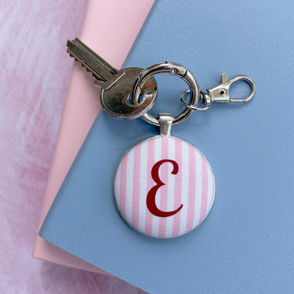 A personalized keychain with a round pink and white striped charm featuring a burgundy monogram letter 'E', attached to a silver snap ring with keys and a lobster clasp, resting on a blue and pink background.