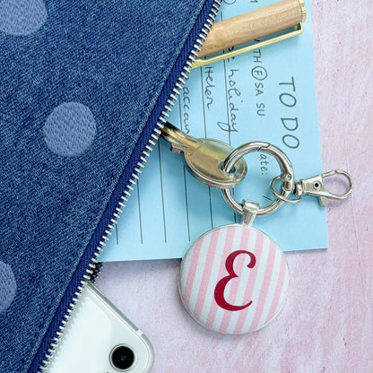 A close-up flat lay showing a blue polka dot zipper pouch partially unzipped to reveal a light blue notepad, gold pen, and a round pink-and-white striped keychain charm with a burgundy letter 'E' monogram, placed on a pink textured surface next to a white phone.