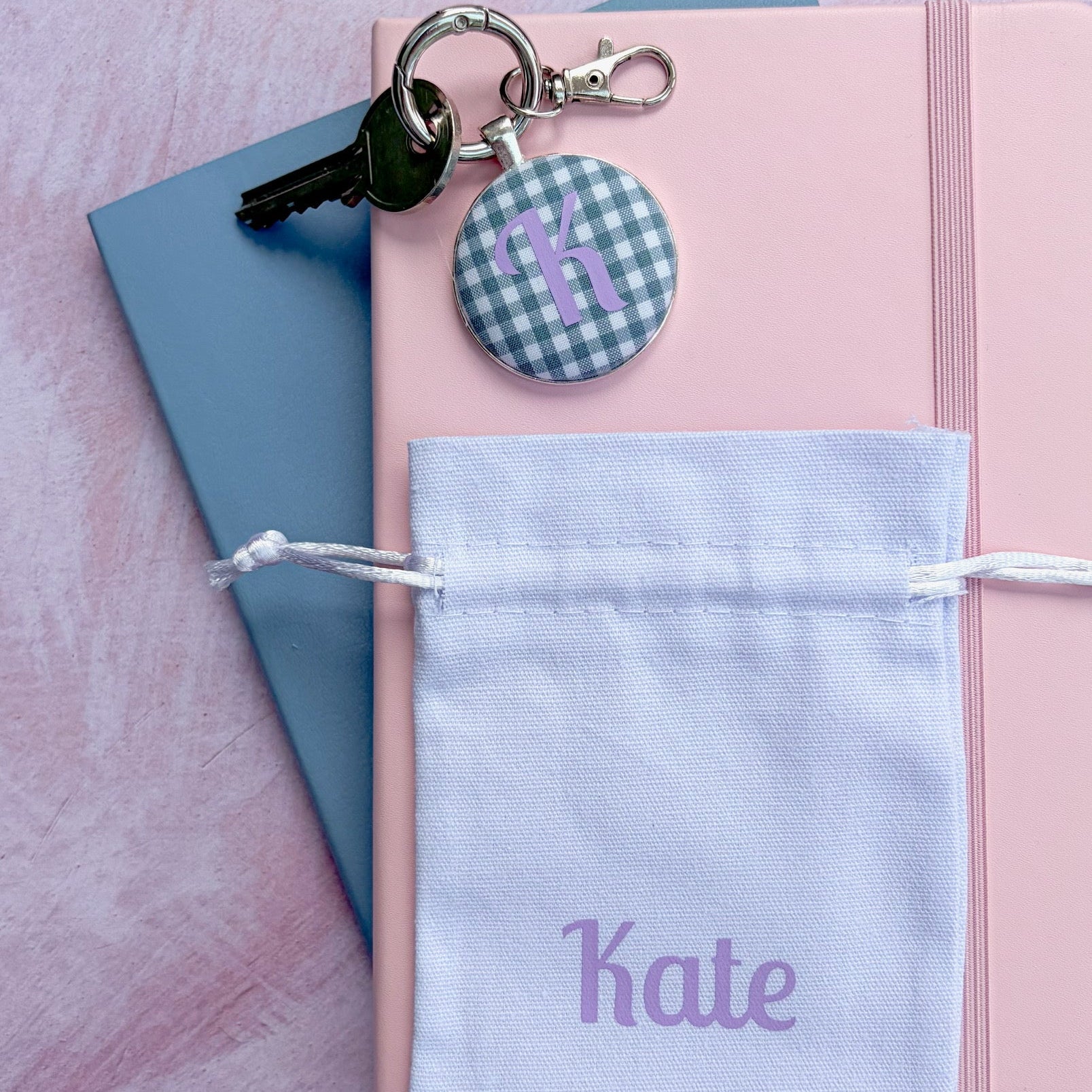 A personalized keychain set featuring a round gingham button with the name 'Kate' in purple, attached to a silver key ring and key, resting on a pink notebook with a white drawstring pouch below.