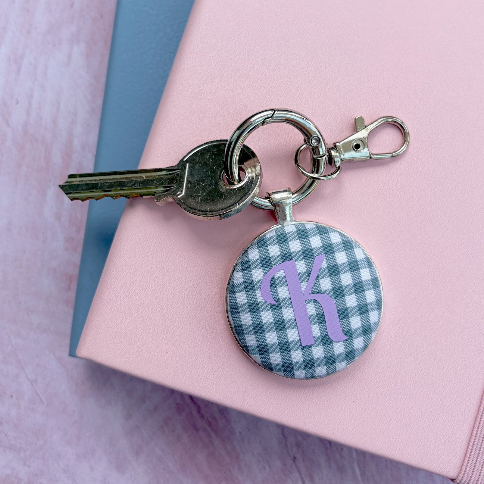 A silver metal keyring with a key and a round charm featuring a green and white gingham check pattern with a purple monogram letter 'k', displayed on a pink notebook.
