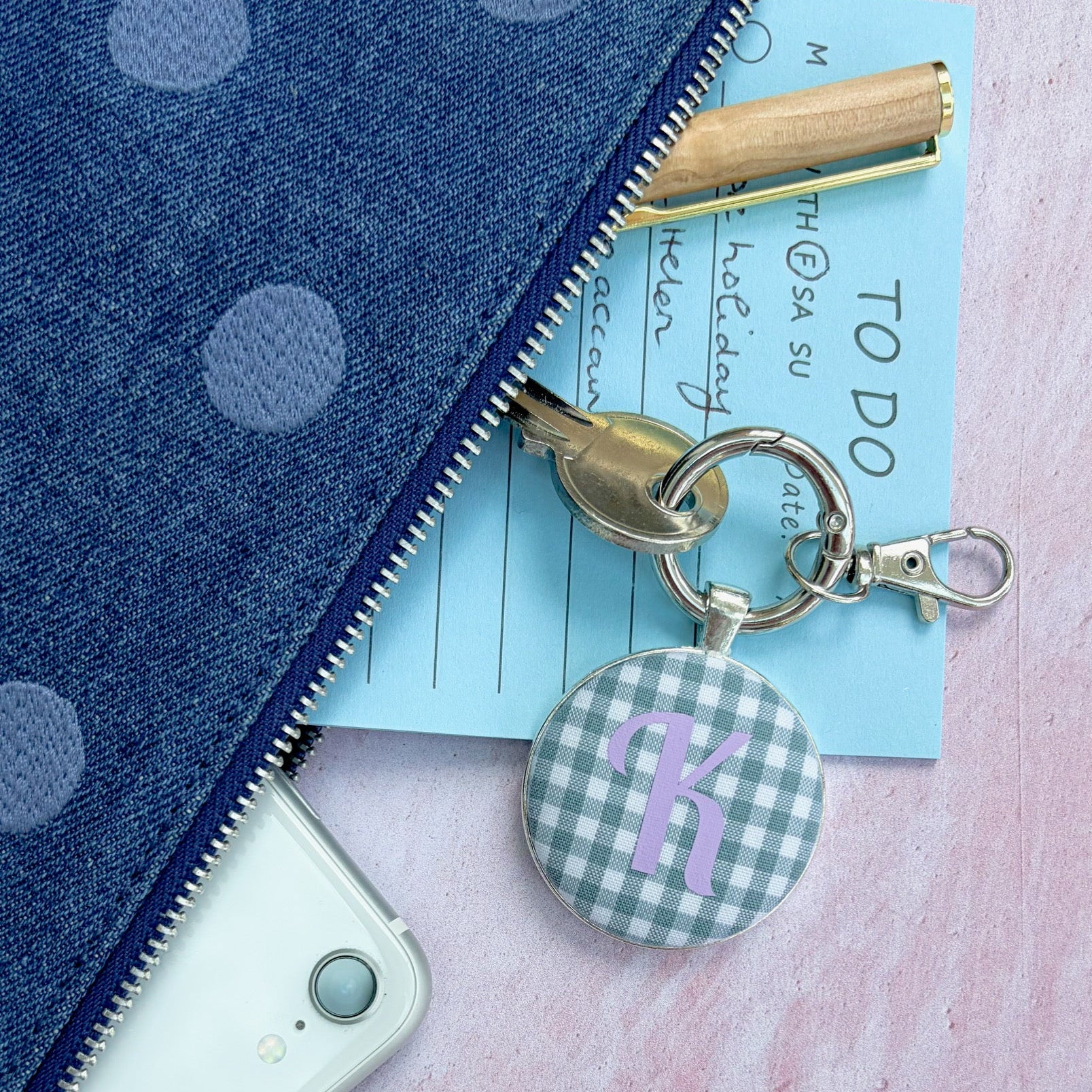 A close-up flat lay of a blue polka dot zip pouch with a gold zipper, shown alongside a white phone, a light blue notepad, a gold pen, and a round gingham keychain charm with a purple letter 'R'.