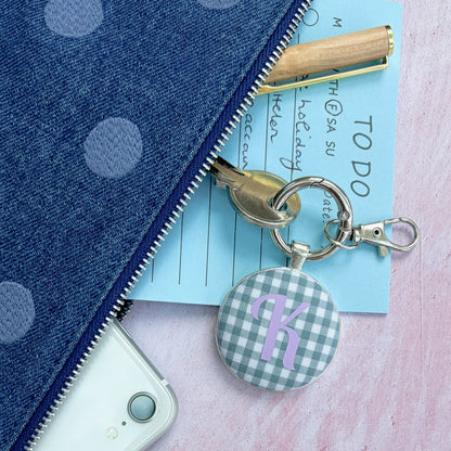 A close-up flat lay of a blue polka dot zip pouch with a gold zipper, shown alongside a white phone, a light blue notepad, a gold pen, and a round gingham keychain charm with a purple letter 'R'.