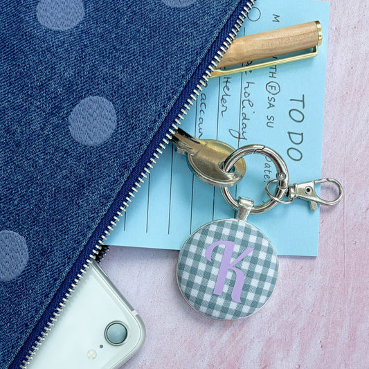 A close-up flat lay of a blue polka dot zip pouch with a gold zipper, shown alongside a white phone, a light blue notepad, a gold pen, and a round gingham keychain charm with a purple letter 'R'.