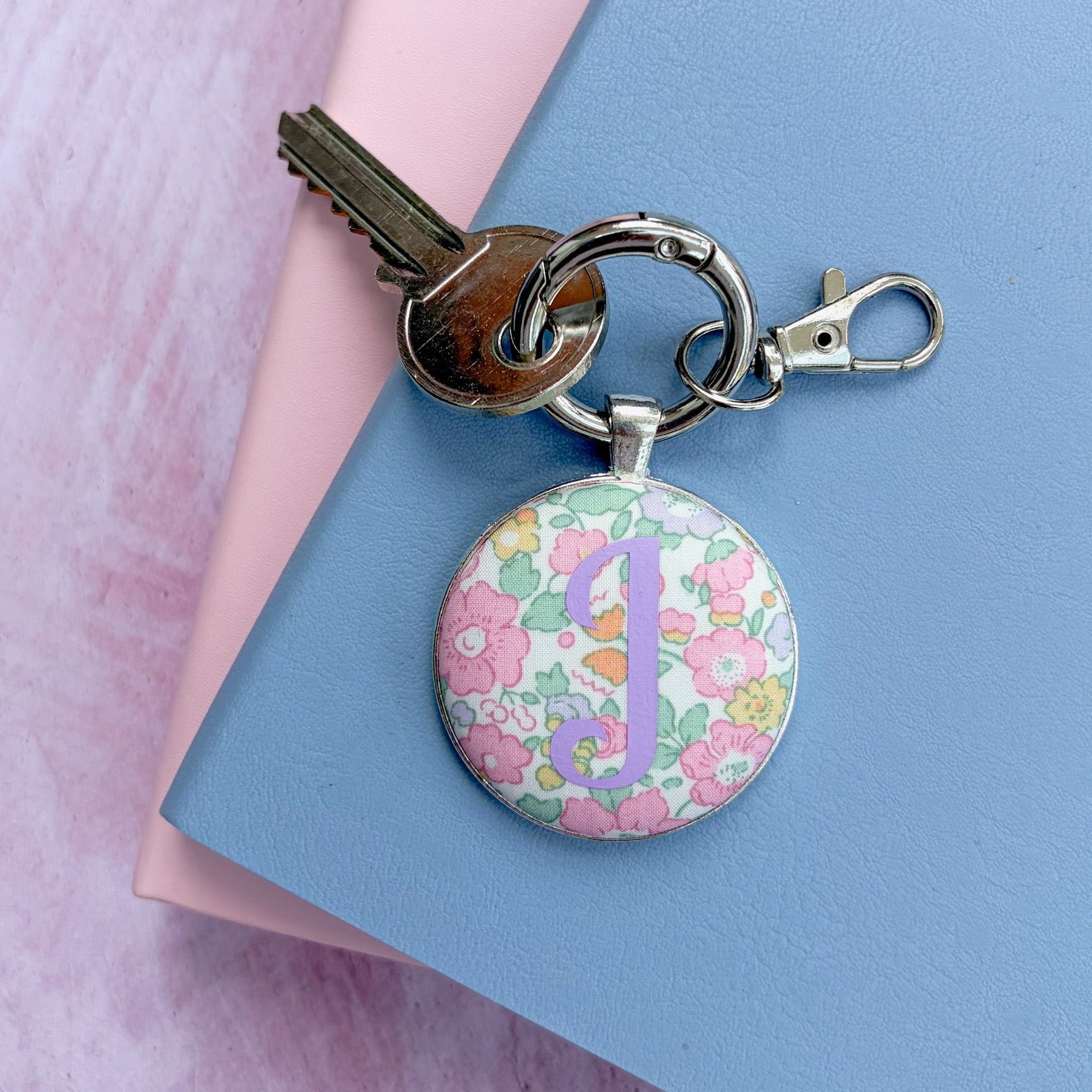 Liberty floral fabric keyring with a lilac personalised initial, on a silver keyring with key and styled on a blue and pink notebook