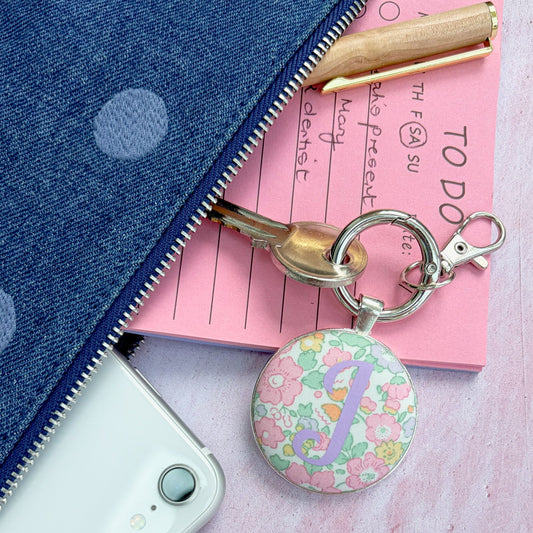 A pastel floral round keyring with a silver key ring and clasp, styled on a pink paper notepad next to a blue polka-dot zipper pouch and a white smartphone.