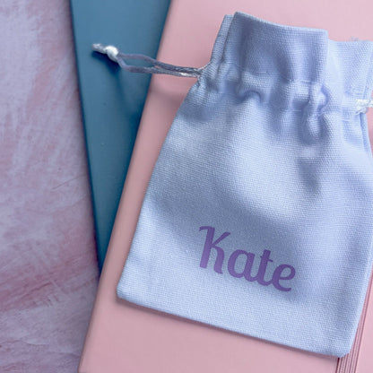 A light blue personalized drawstring pouch printed with the name 'Kate' in purple, resting on top of a pink notebook and a teal book on a textured pink surface.