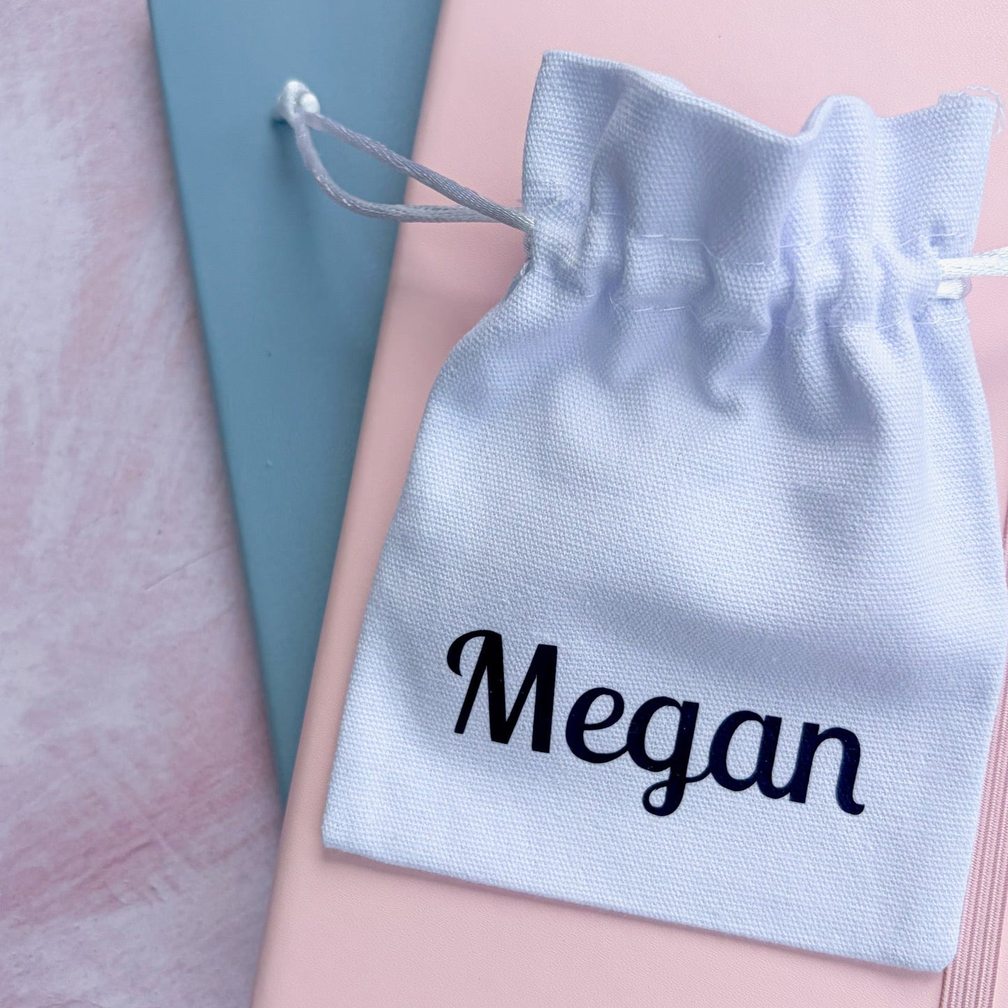 A white personalized drawstring pouch printed with the name 'Megan' in navy script, resting on top of a pink notebook and a blue book on a pink textured surface.