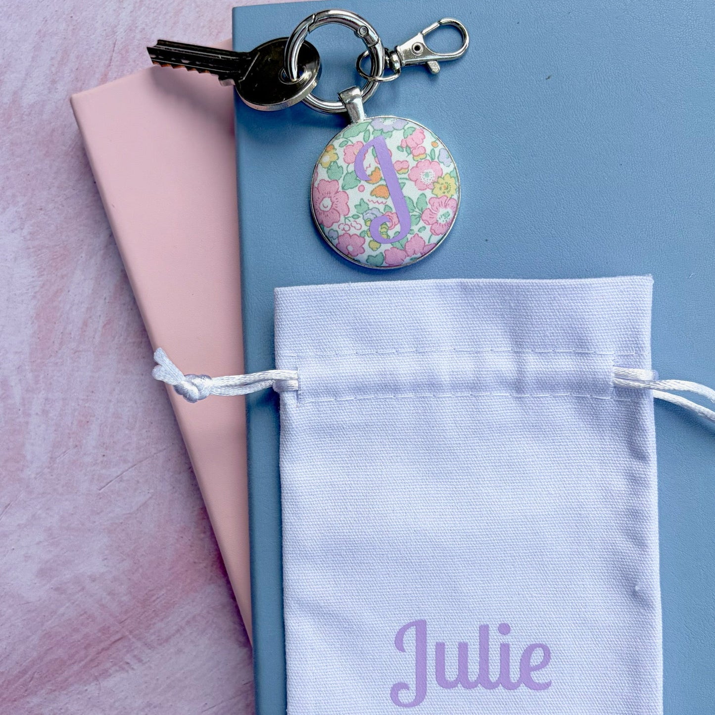 A personalized keychain with a round floral button charm and a silver clasp, displayed on a light blue notebook with a pink folder underneath. A white drawstring pouch printed with the name 'Julie' in purple is in the foreground, all set against a pink textured background.