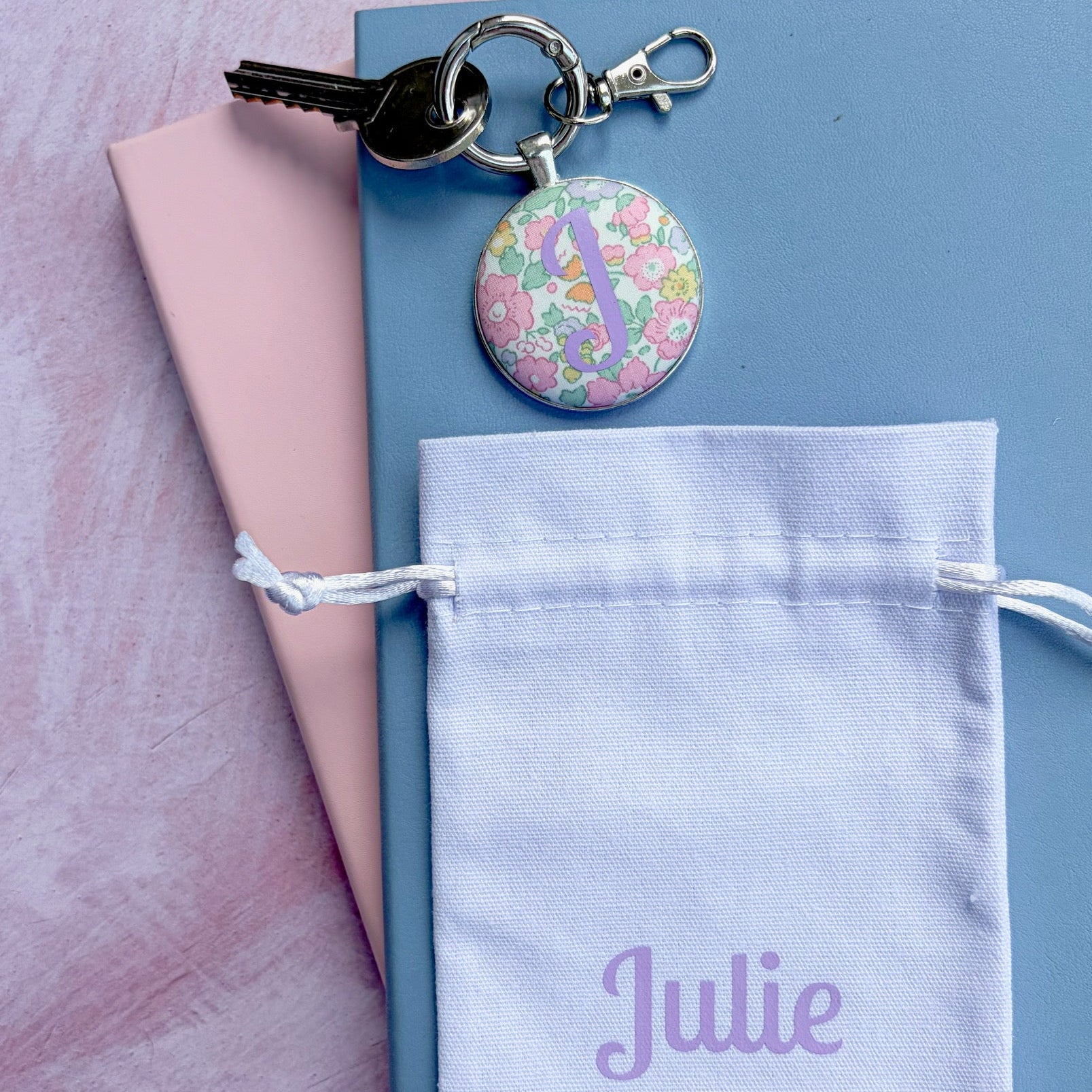 A personalized keychain with a round floral button charm and a silver clasp, displayed on a light blue notebook with a pink folder underneath. A white drawstring pouch printed with the name 'Julie' in purple is in the foreground, all set against a pink textured background.