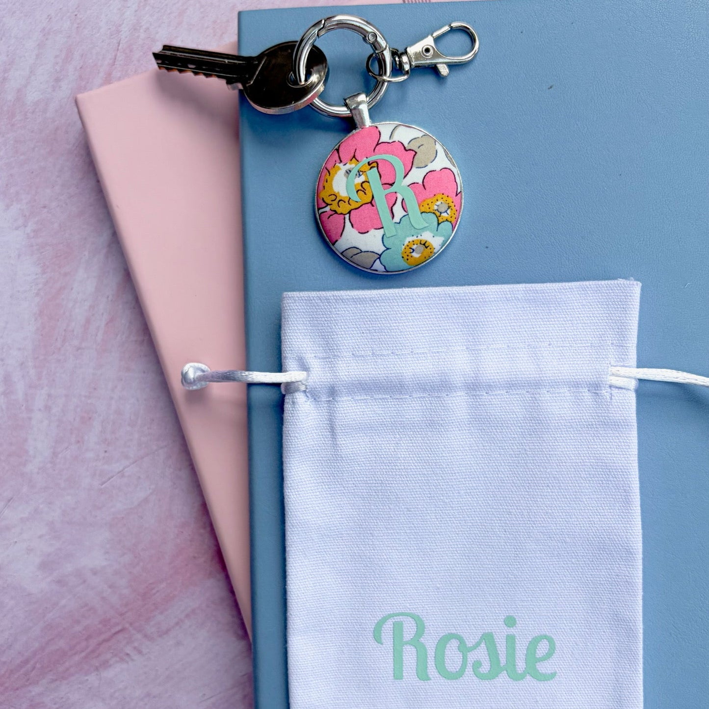A personalized keyring with a round floral charm and a key, displayed on top of a blue notebook with a pink page, next to a white drawstring pouch printed with the name 'Rosie' in mint green, all on a pink textured background.